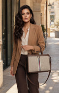 A woman in a tan blazer and brown trousers carries a structured tote bag with a beige monogram pattern and a brown leather trim and strap, set against a blurred outdoor urban background.